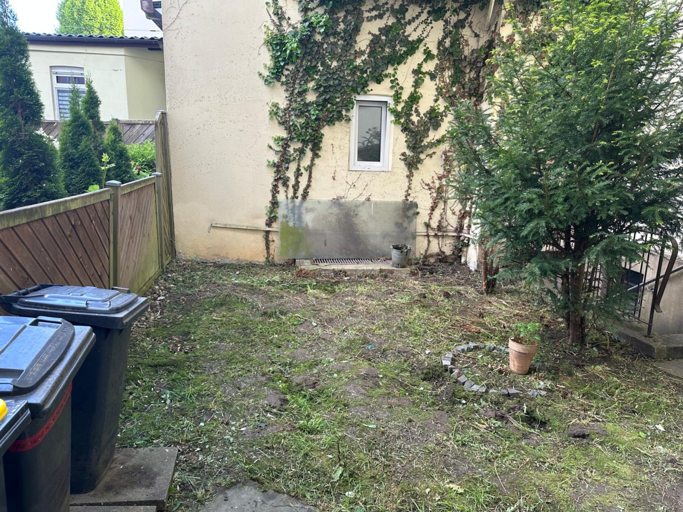 Hinterhof Nachher Bild Dortmund Ein leerer Garten mit Mülltonnen, einer Wand und einigen Bäumen im Hintergrund.