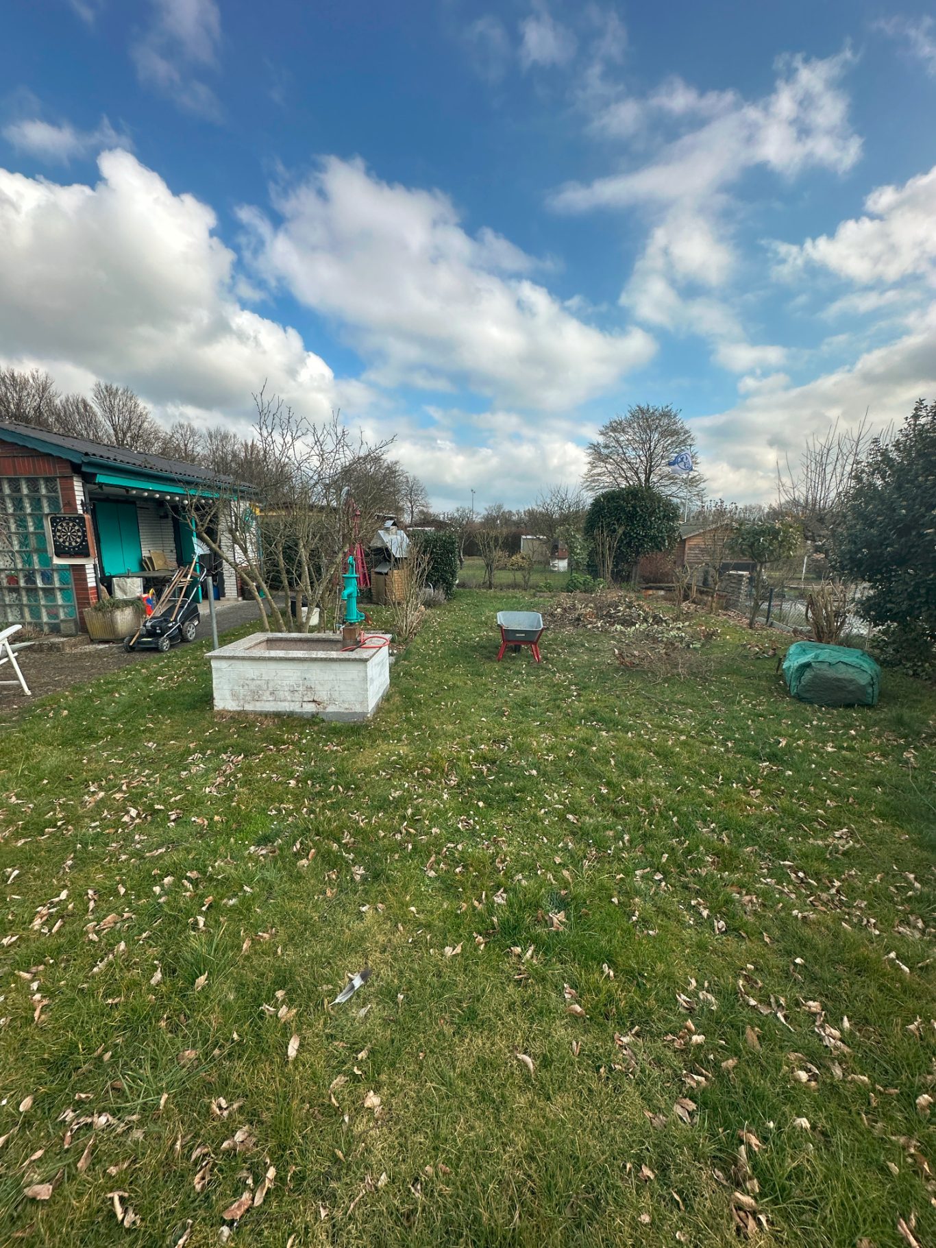 Vorher Bild eines Gartens vor unseren Dienstleistungen Gemüsegarten mit Beeten, Bäumen und bunten Hütten unter bewölktem Himmel.