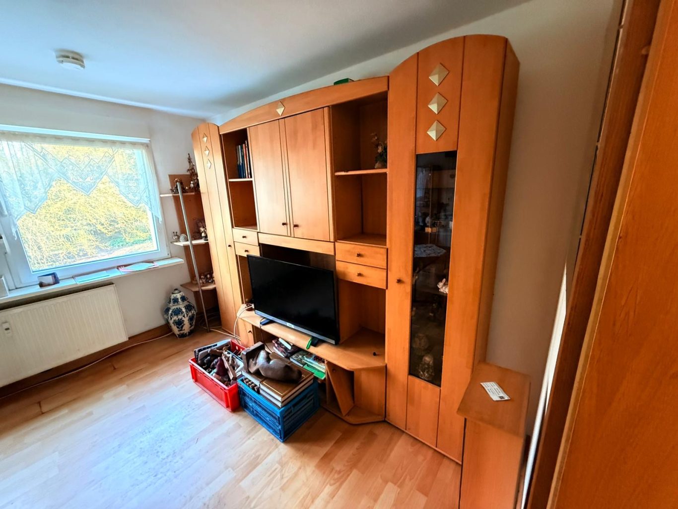 Vorher Bild Wohnungsauflösung Wohnzimmer mit Holzschrank, Fernseher und Körben auf dem Boden.