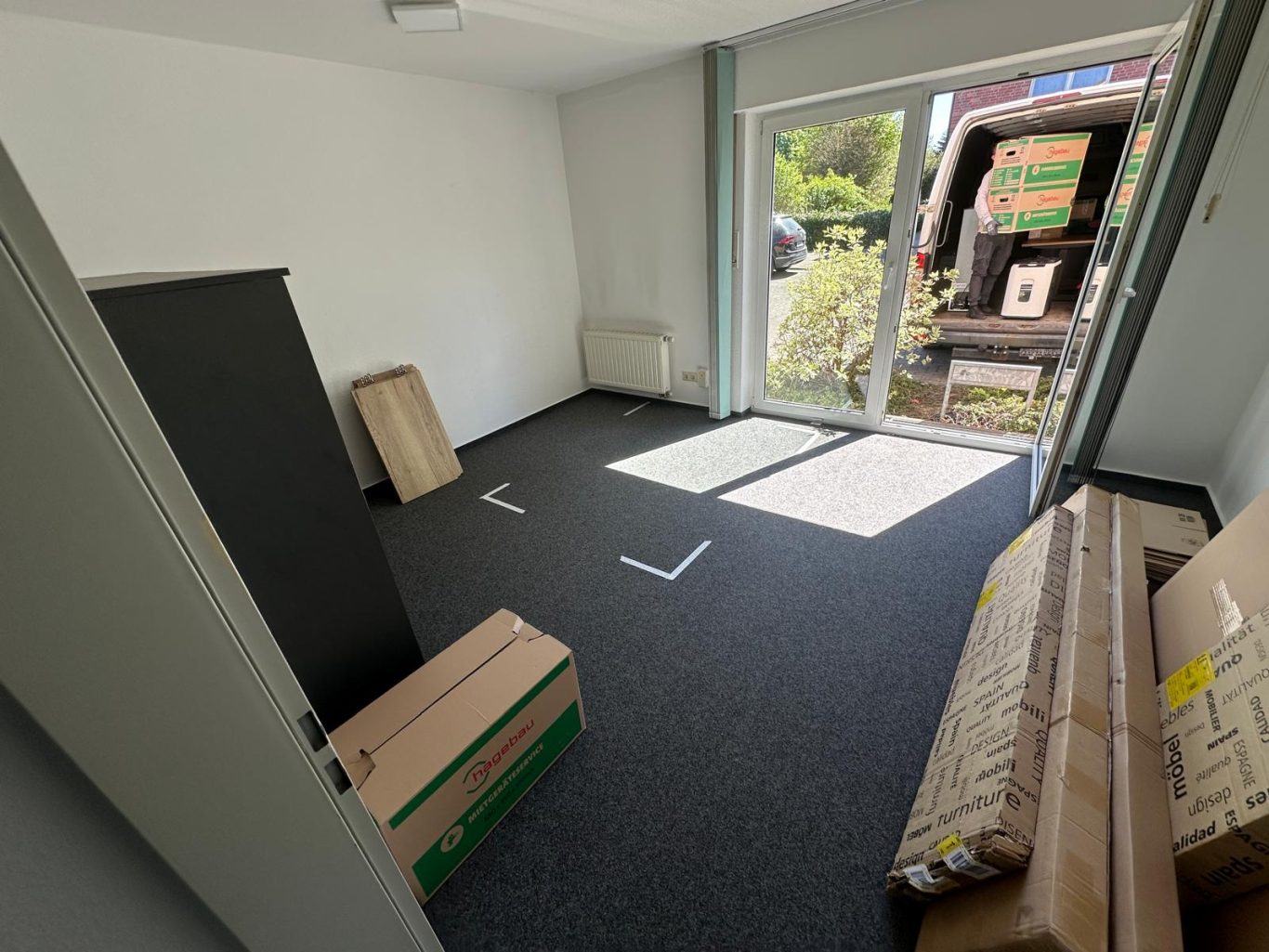 Vorherbild Büroraum vor Umzug Leerer Raum mit dunklem Boden, Fenster und Umzugskartons im Hintergrund.