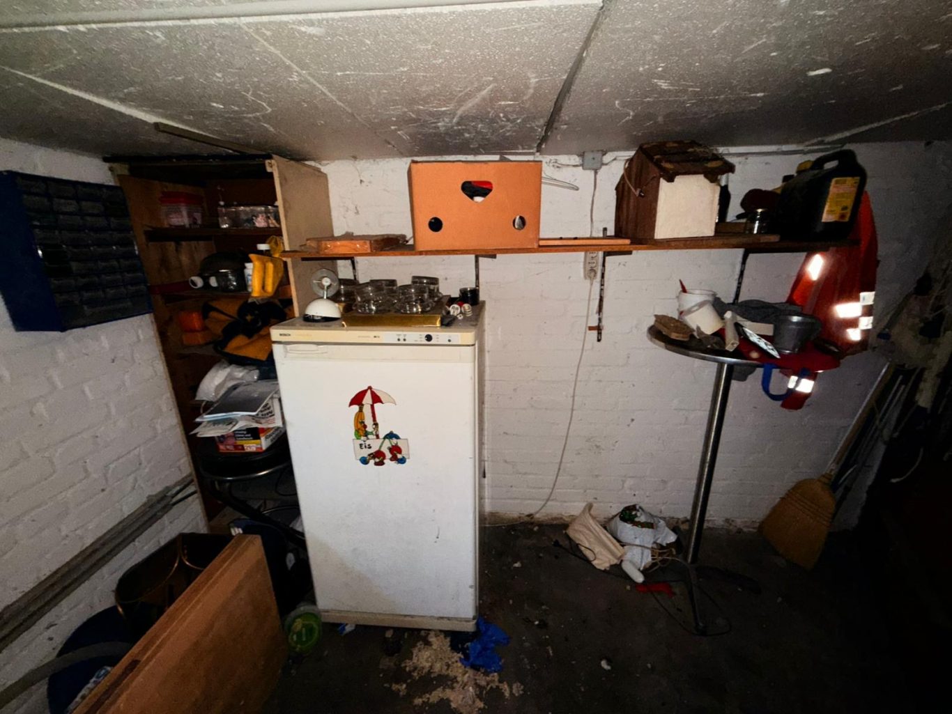 Vorher Bild Keller Entrümpelung Kellerraum mit einem Kühlschrank, Regalen und verschiedenen Gegenständen.
