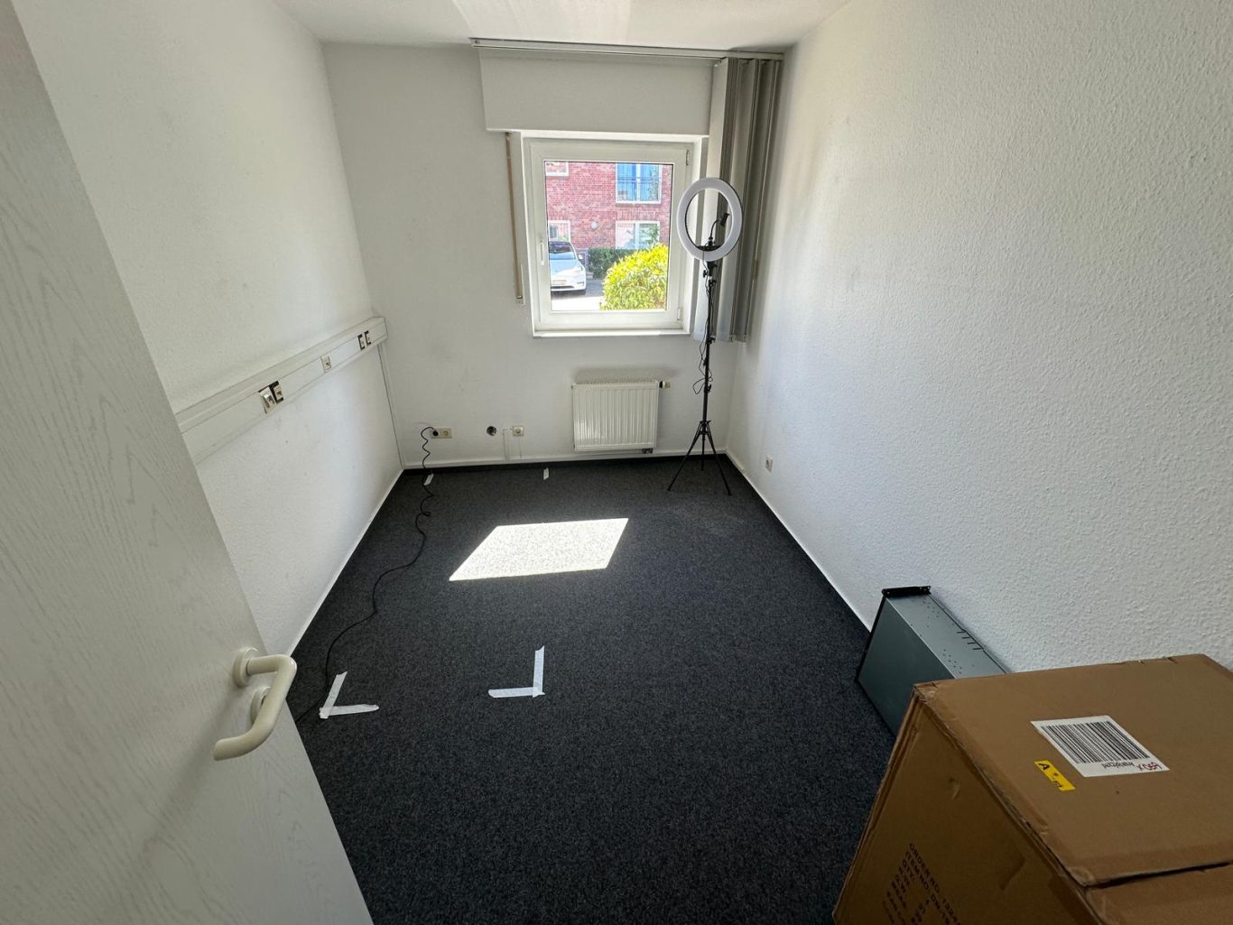 Vorher Bild Büroumzug Kleine, leere Zimmer mit einem Fenster, Teppichboden und einer Kiste in der Ecke.